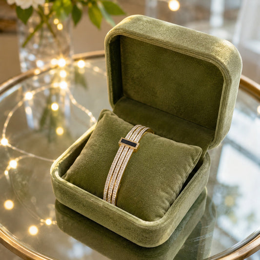 A gold-toned multi-strand bracelet with a black rectangular center accent, displayed inside an open olive green velvet jewelry box 