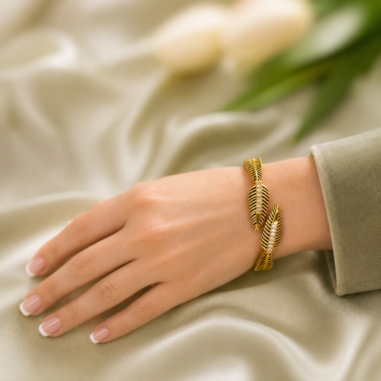 Gold feather cuff bracelet with crystal accents worn on the wrist against a soft sage satin background
