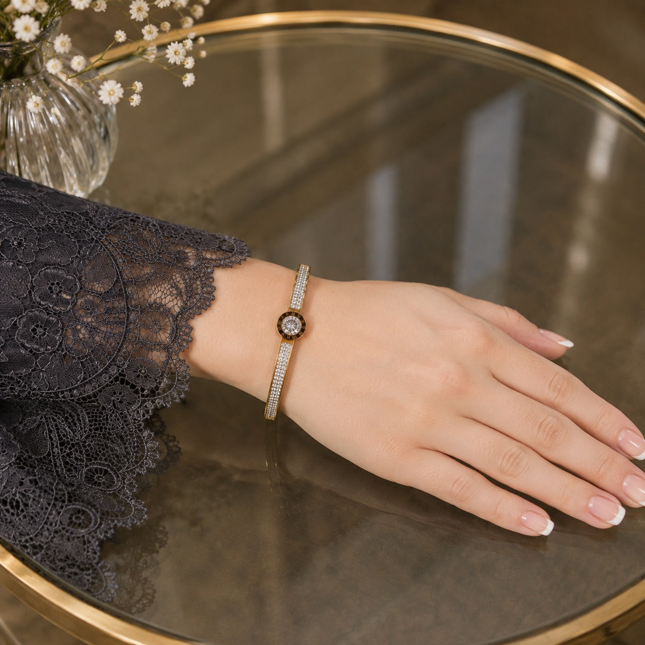Gold crystal lock bracelet worn on a hand resting on a glass-top table with a black lace sleeve and soft floral background.