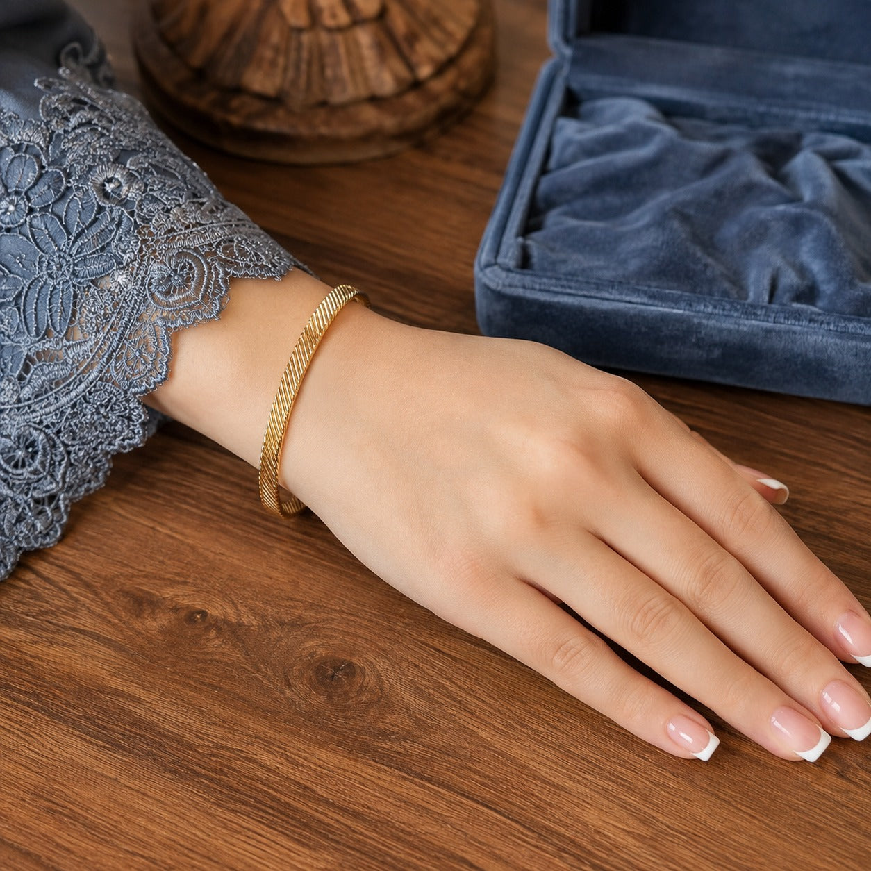 Gold twisted open cuff bracelet worn on a hand with a smoky blue lace sleeve resting on a walnut wood table beside a blue velvet jewelry box.