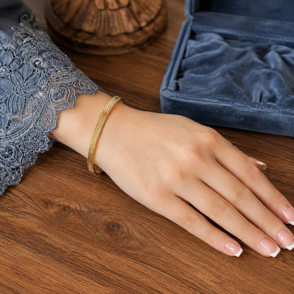 Gold twisted open cuff bracelet worn on a hand with a smoky blue lace sleeve resting on a walnut wood table beside a blue velvet jewelry box.