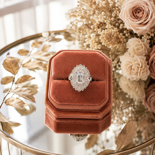 S925 sterling silver halo statement ring with a radiant-cut center stone displayed in a terracotta velvet octagonal ring box on a reflective glass table with floral décor.