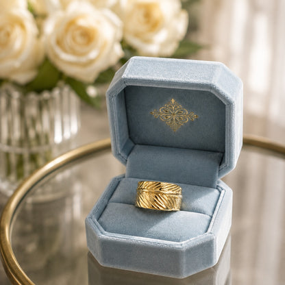 Wide gold leaf-textured band ring displayed in a soft powder blue velvet ring box on a reflective glass tabletop with blurred white roses.