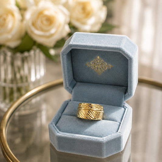Wide gold leaf-textured band ring displayed in a soft powder blue velvet ring box on a reflective glass tabletop with blurred white roses.
