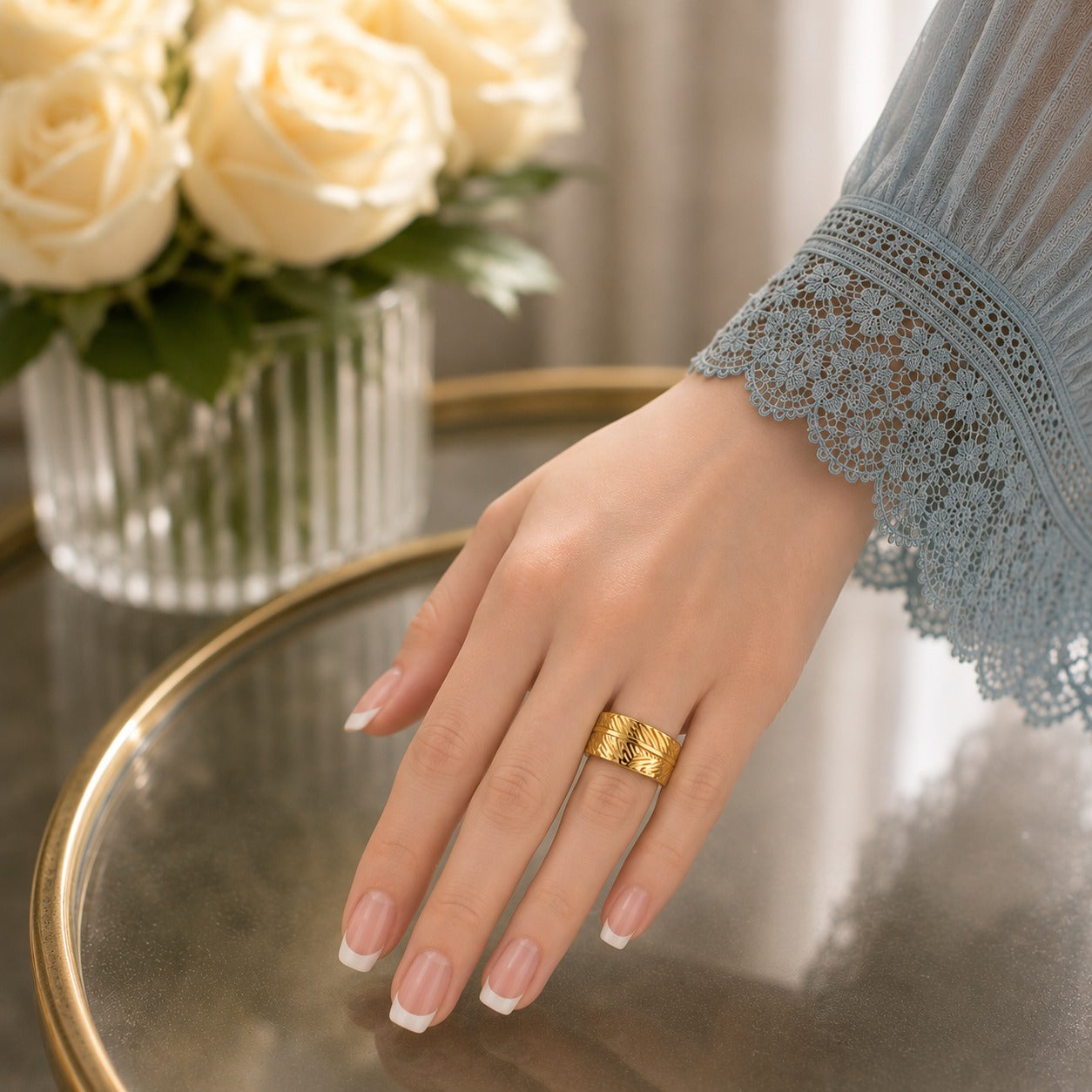 Woman wearing a wide gold botanical leaf-pattern band ring with a soft blue lace sleeve, resting her hand on a reflective glass surface beside white roses.
