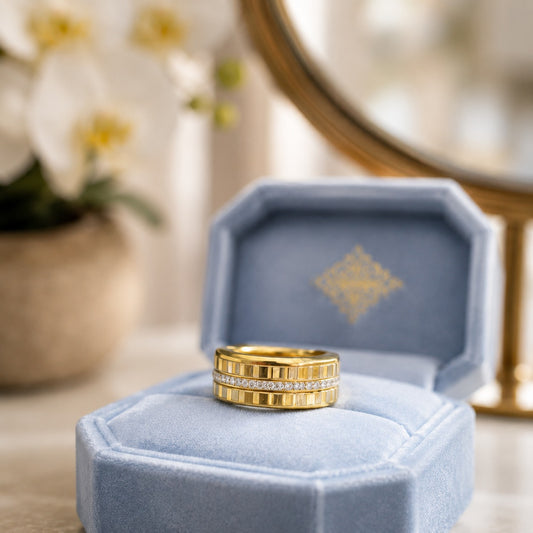 Gold-tone stainless steel crystal band showcased inside an open powder blue velvet ring box beside white orchids and a decorative vanity mirror.