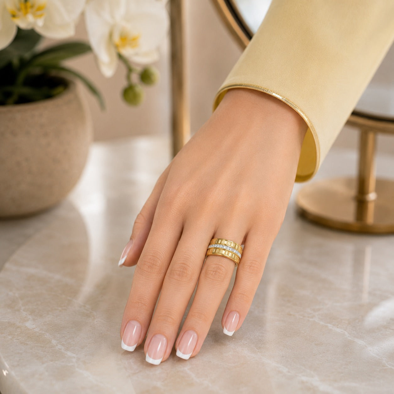 Gold-tone stainless steel ridged band with a crystal center strip worn on a hand against a polished marble vanity with soft floral décor.