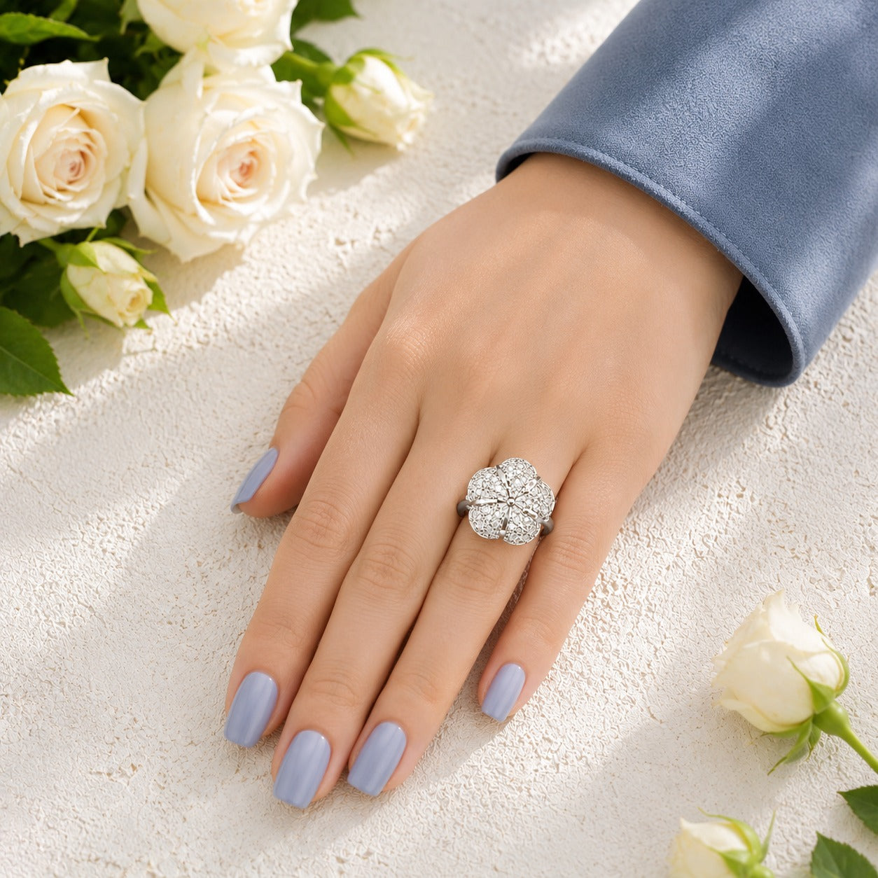Floral crystal statement ring worn on a hand with soft blue manicure and white rose background.
