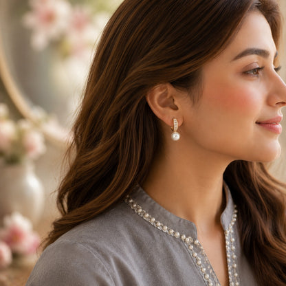 A close-up side profile of a woman with long brown hair wearing a silver-toned drop earring with a white pearl, paired with a gray embroidered top.