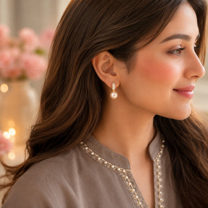 A close-up side profile of a woman with long brown hair wearing a delicate gold and pearl drop earring, paired with a taupe embroidered kurta. The background is a soft, blurred indoor setting with pink flowers and warm bokeh lights.