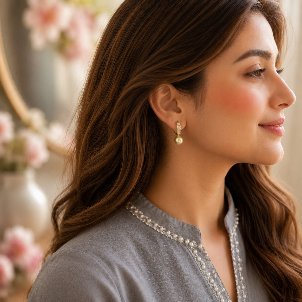A woman in profile wearing a delicate gold and pearl drop earring, styled with a gray embroidered top against a softly blurred background with pink flowers.