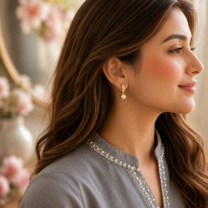 A woman in profile wearing a delicate gold and pearl drop earring, styled with a gray embroidered top against a softly blurred background with pink flowers.