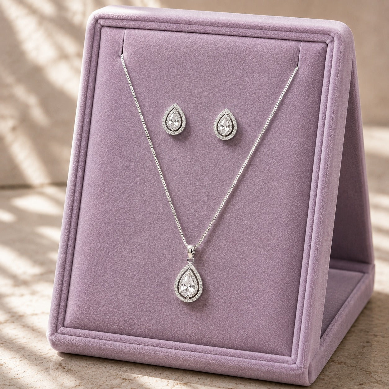 Clear zircon teardrop pendant necklace and matching stud earrings displayed on a soft lilac jewellery stand.