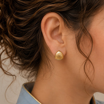 A close-up of a model's ear wearing a small, teardrop-shaped gold stud earring with a textured, ribbed surface.