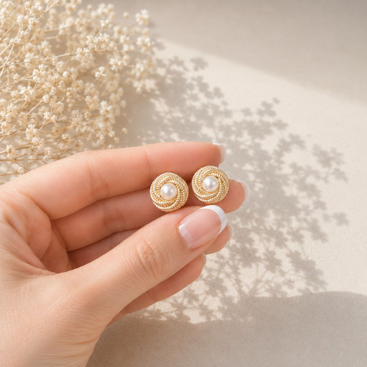 Gold-tone round stud earrings with a creamy pearl centre surrounded by layered rope-textured circular coils held in a hand