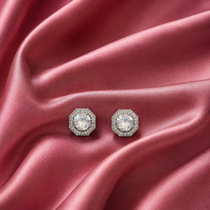 Pair of silver-tone geometric stud earrings with clear faceted centre zircons and detailed zircon-set borders displayed on rose pink satin.