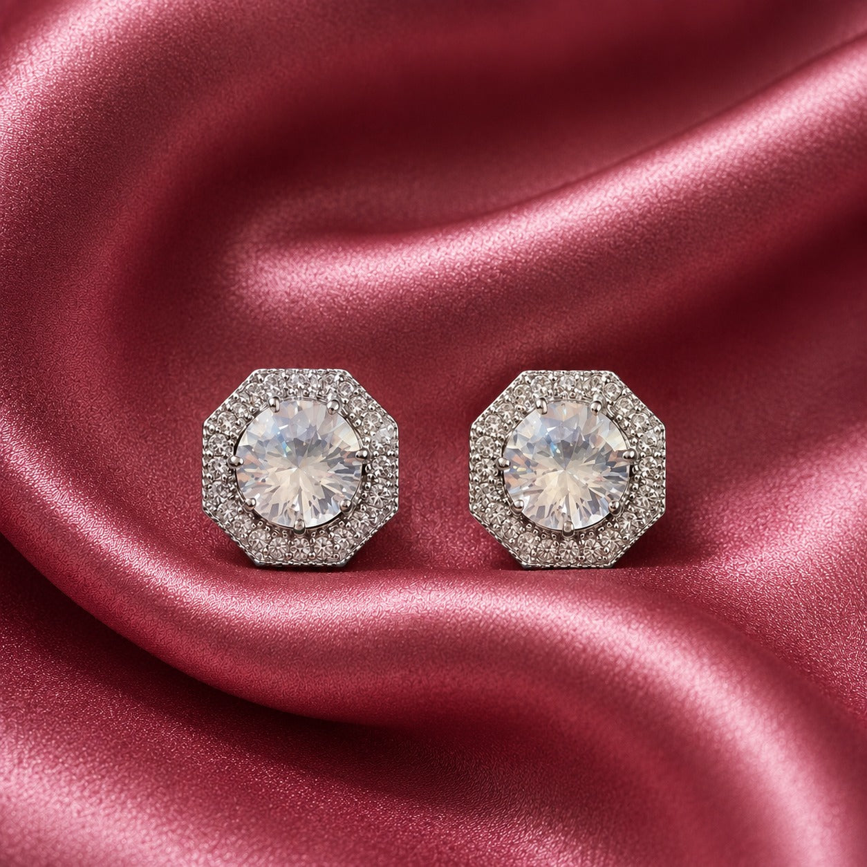 Pair of silver-tone geometric stud earrings with clear faceted centre zircons and detailed zircon-set borders displayed on rose pink satin.