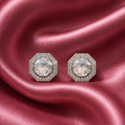 Pair of silver-tone geometric stud earrings with clear faceted centre zircons and detailed zircon-set borders displayed on rose pink satin.