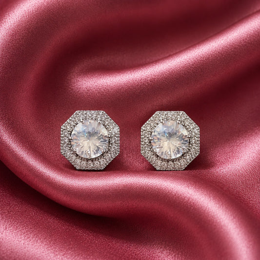 Pair of silver-tone geometric stud earrings with clear faceted centre zircons and detailed zircon-set borders displayed on rose pink satin.
