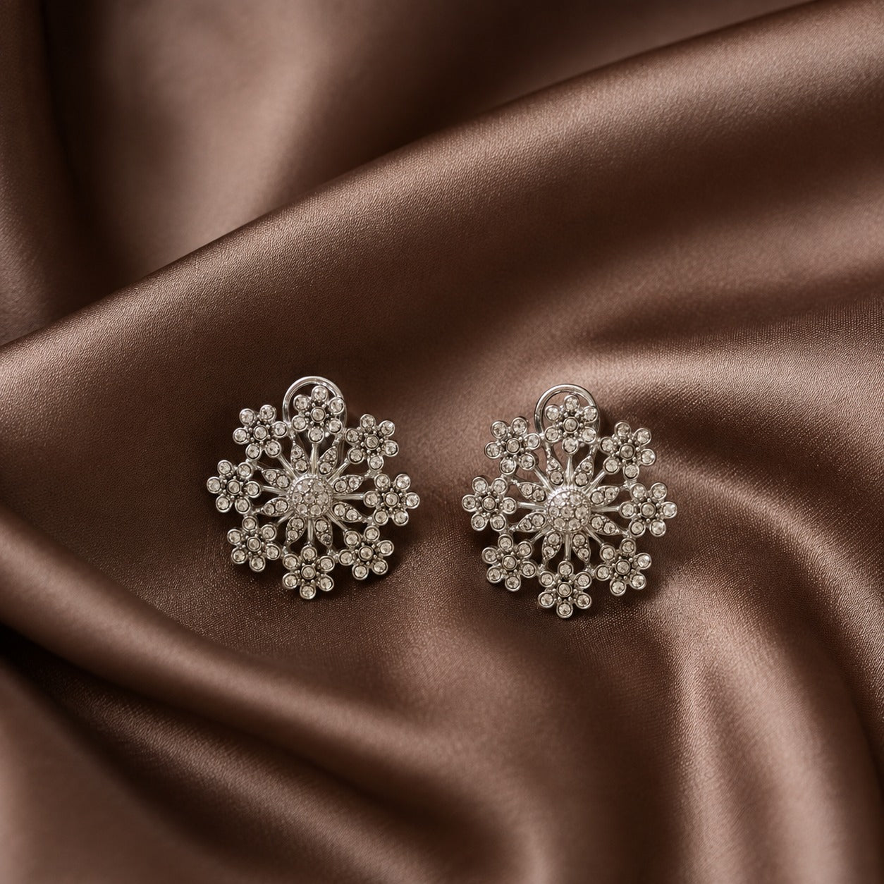 Pair of silver-tone floral statement earrings with intricate clear crystal clusters and openwork detailing displayed on brown satin.