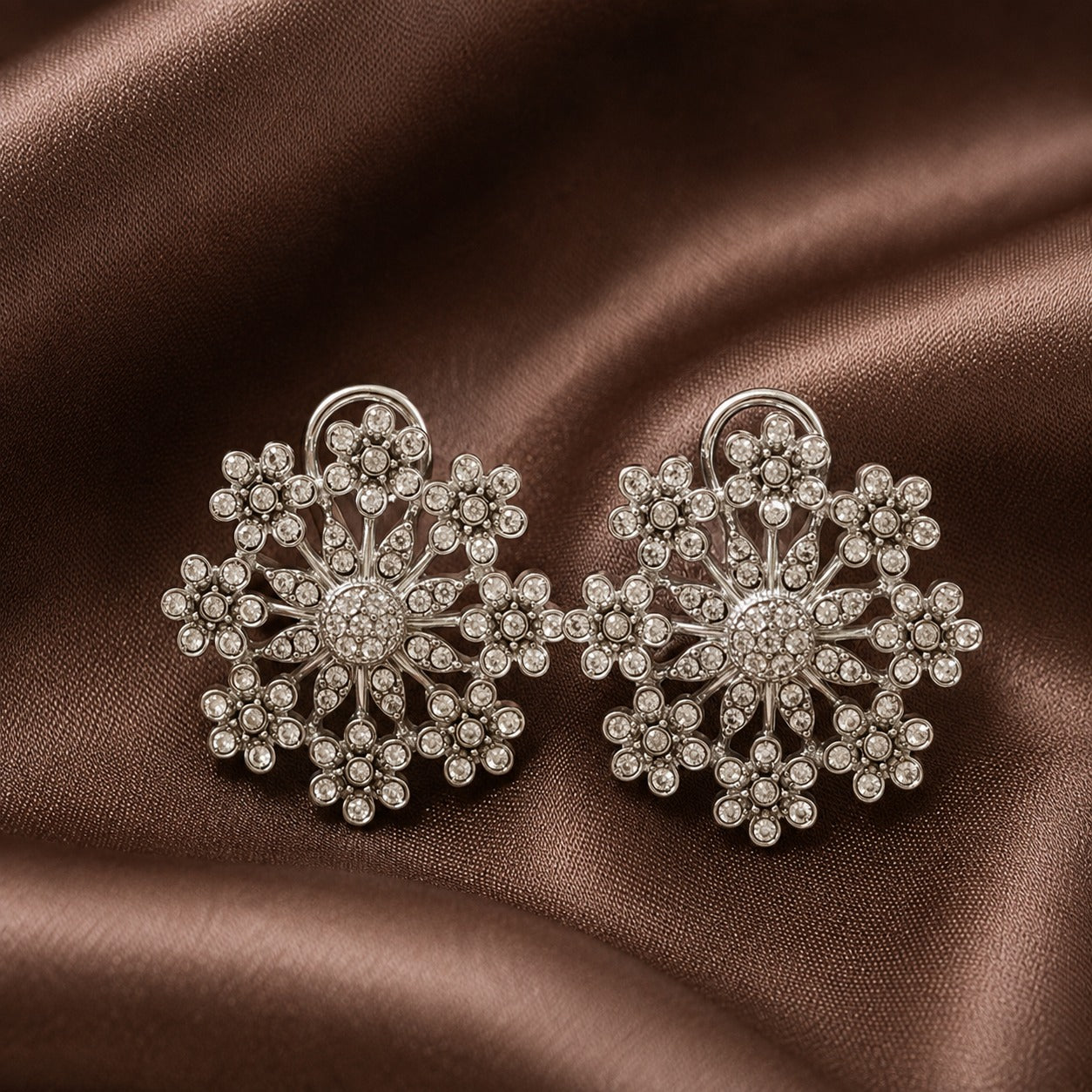 Pair of silver-tone floral statement earrings with intricate clear crystal clusters and openwork detailing displayed on brown satin.