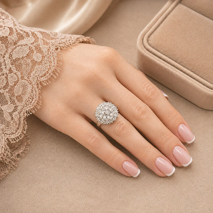 A close-up of a hand wearing a sparkling round cluster ring with clear stones, styled with a beige lace sleeve and a matching jewelry box in the background.