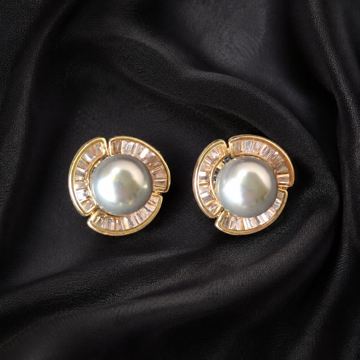 Gold-tone Pearl Circuit Stud Earrings with luminous round pearl centres and segmented baguette-cut clear stone frames displayed on black fabric.