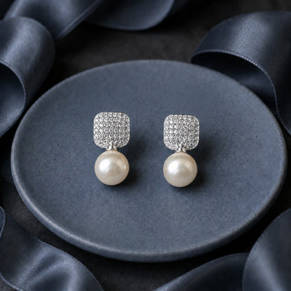Pearl Grid Drop Earrings featuring rounded-square silver-tone tops with tiny clear stones and luminous white pearl drops, displayed on a dark grey surface.