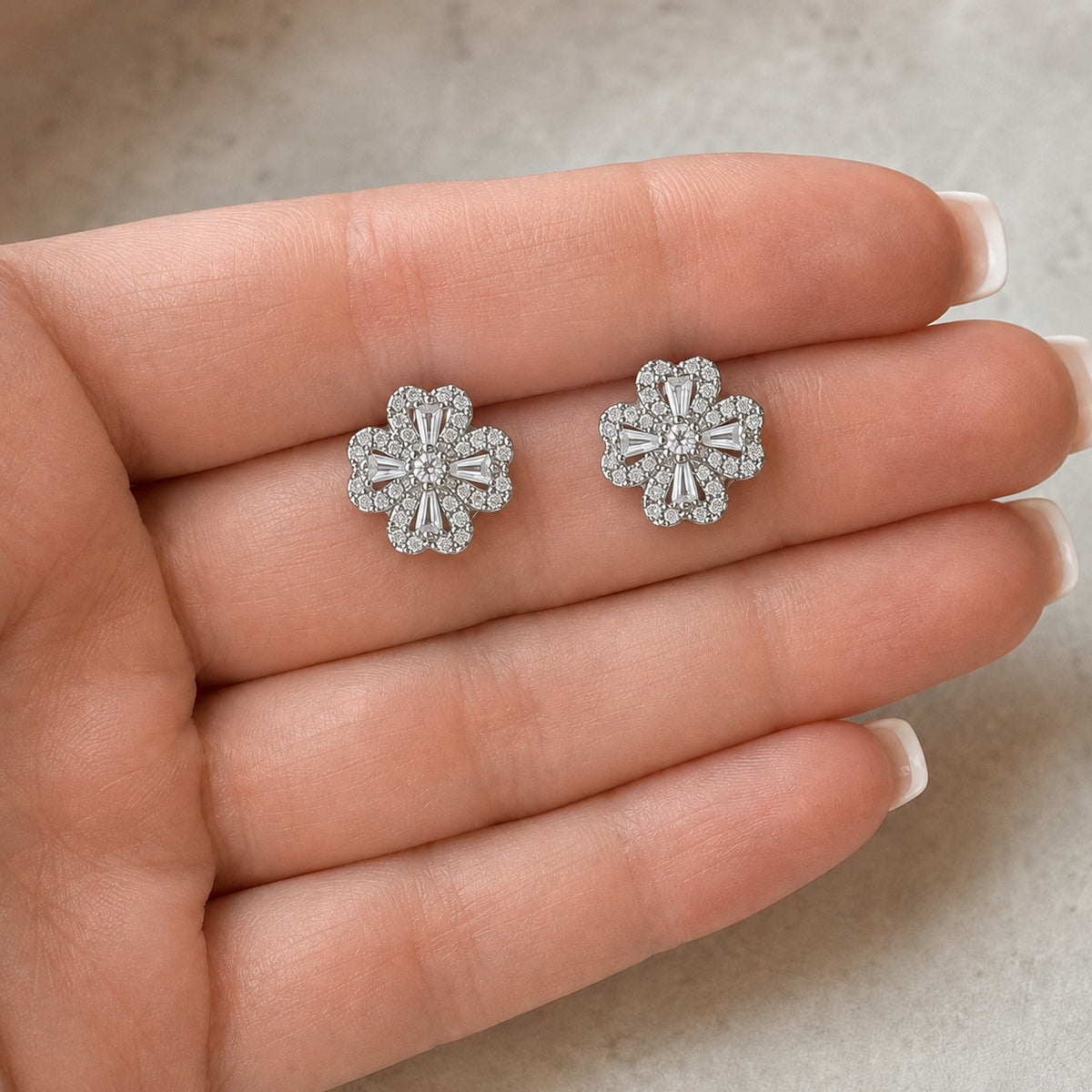 Pair of S925 silver Prism Clover Stud earrings displayed on a hand, showing the detailed scalloped petals, tapered baguette stones and clear zircon centre.