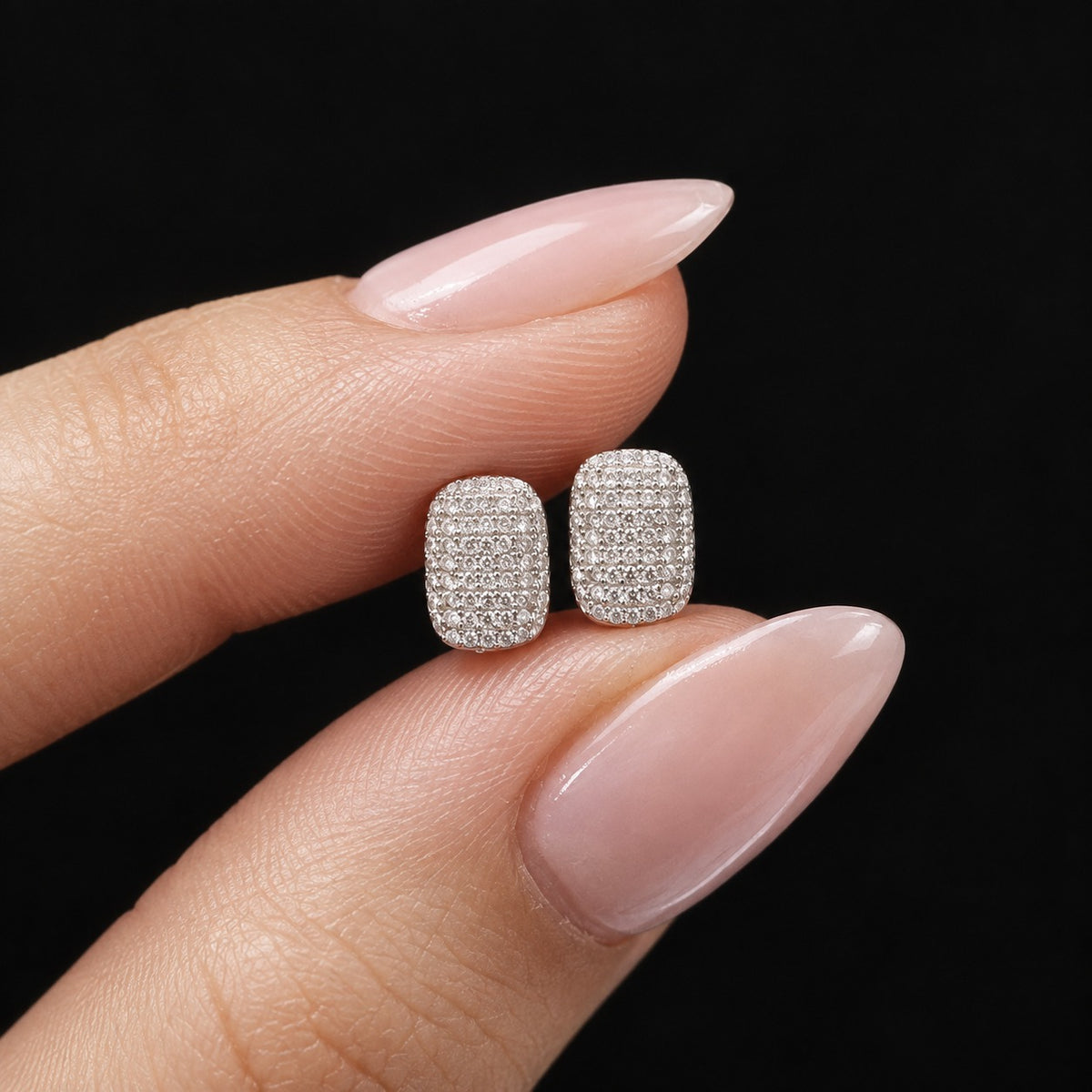 Close-up of small silver rectangular crystal stud earrings held between fingers, showing their rounded corners and detailed stone-set surface.