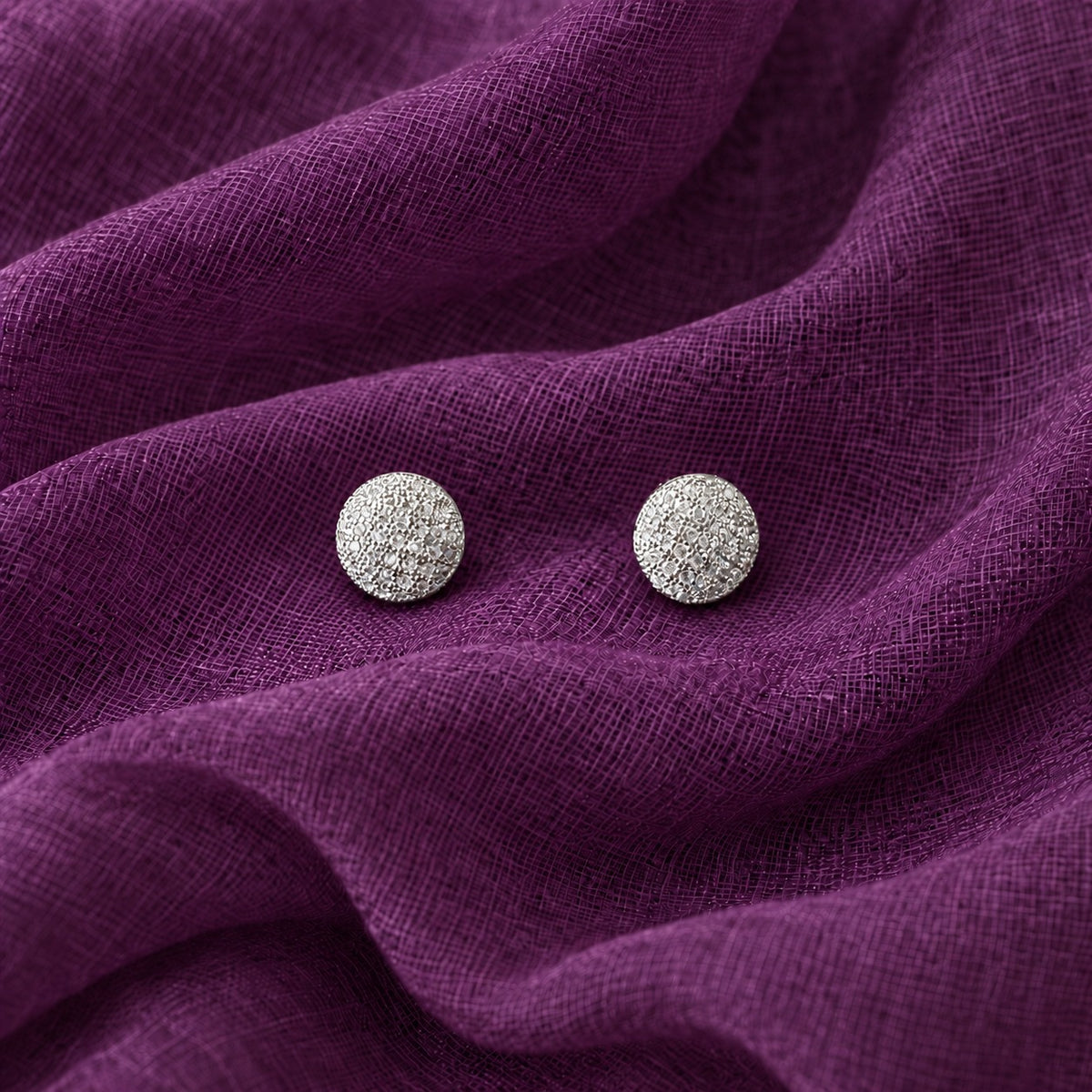 Close-up of round silver-tone stud earrings with intricate clustered clear zircon detailing on purple textured fabric.