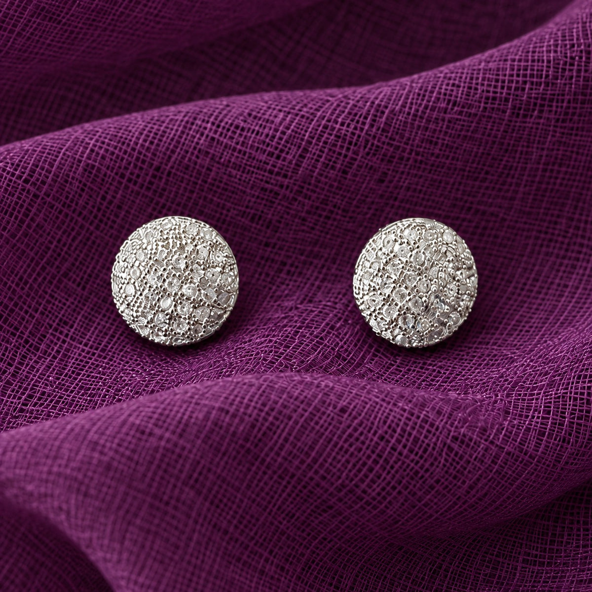 Close-up of round silver-tone stud earrings with intricate clustered clear zircon detailing on purple textured fabric.