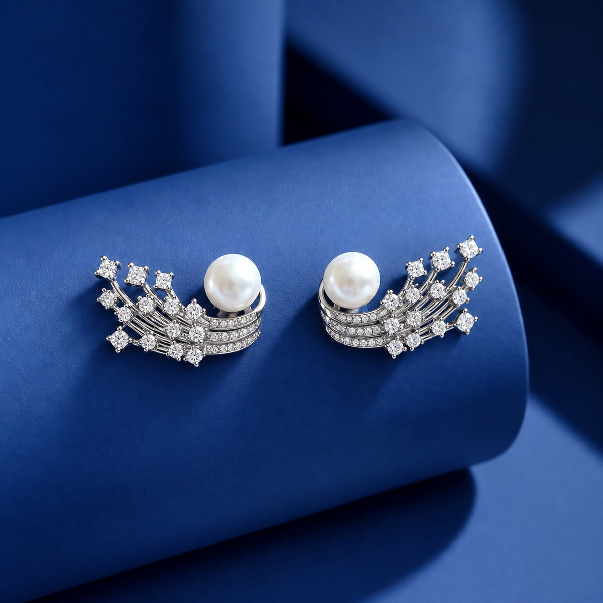 Silver-tone statement earrings with white pearls, curved rows of clear zircon stones and branching crystal details displayed on a blue surface