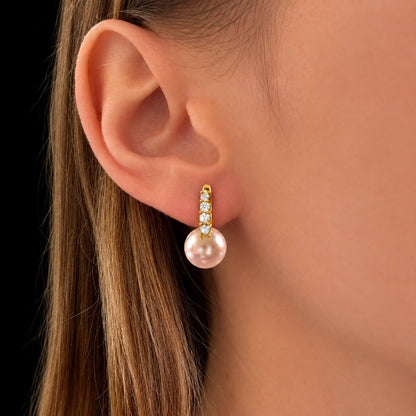 Close-up of a model wearing gold-tone earrings featuring a vertical row of clear stones and a luminous blush-pink pearl drop.