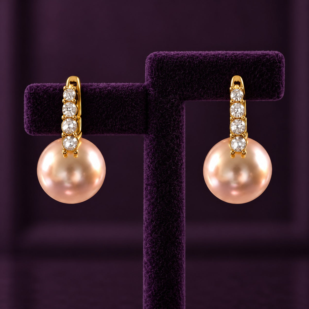 Gold-tone drop earrings with four clear stones and large blush-pink pearl drops displayed on a deep plum jewellery stand.