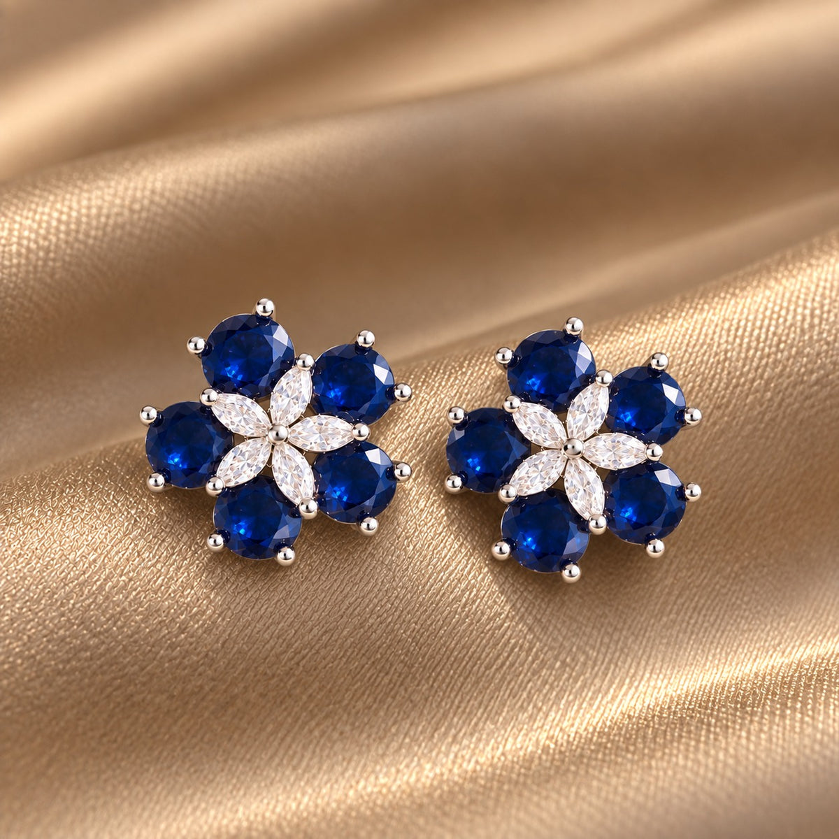 Pair of Fivefold Bloom Studs featuring five faceted  sapphire blue stones surrounding a clear marquise-cut crystal flower in a polished silver-tone setting, displayed on beige satin.