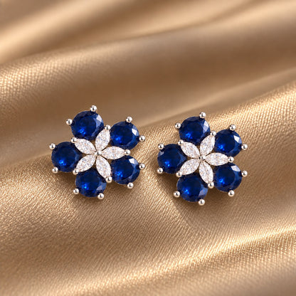 Pair of Fivefold Bloom Studs featuring five faceted  sapphire blue stones surrounding a clear marquise-cut crystal flower in a polished silver-tone setting, displayed on beige satin.