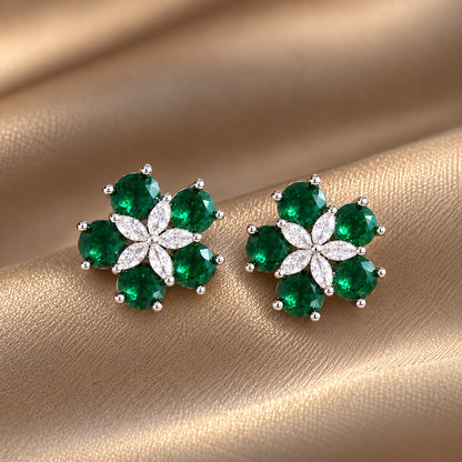 Pair of Fivefold Bloom Studs featuring five faceted  emerald green stones surrounding a clear marquise-cut crystal flower in a polished silver-tone setting, displayed on beige satin.