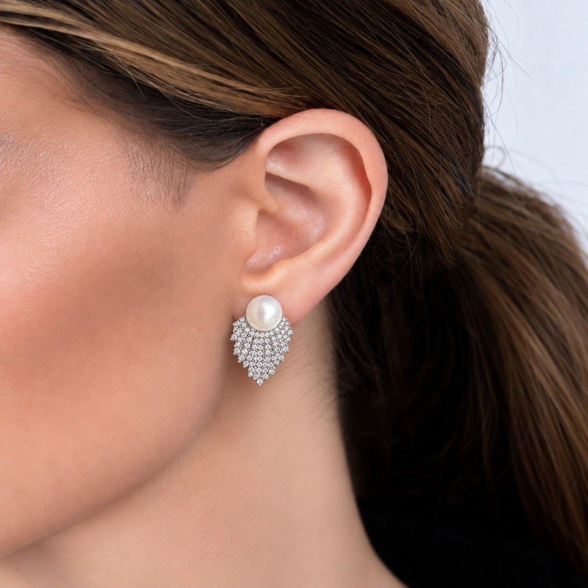 Close-up of a woman wearing Pearl Cascade Studs, featuring a round white pearl above layered silver-tone rows of clear zircon that taper below the earlobe.