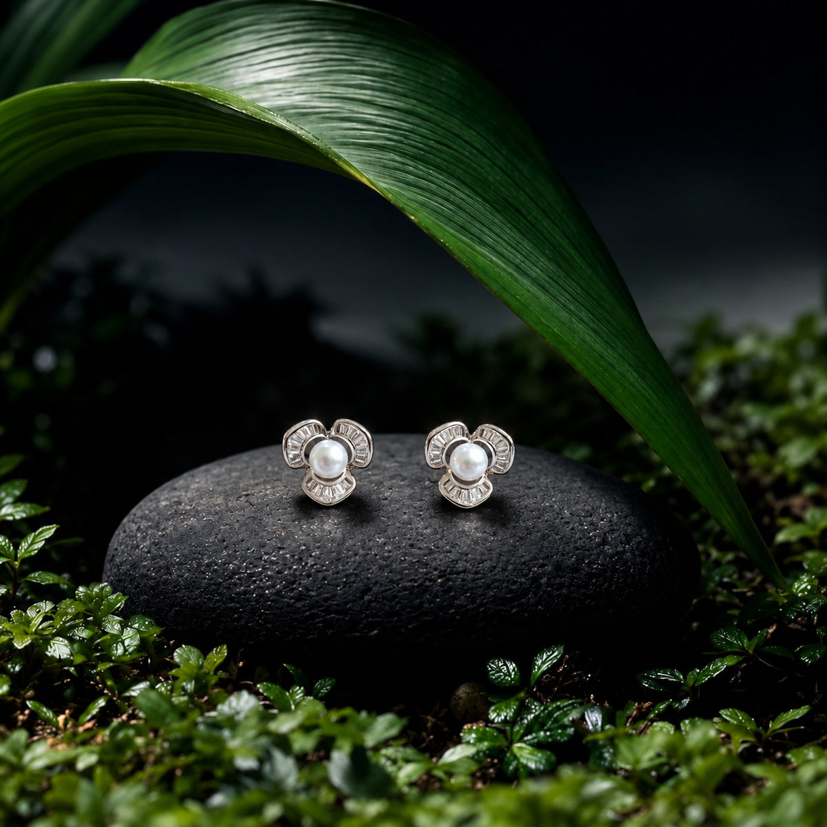 “Pair of silver-tone Pearl Arc Stud Earrings displayed on a black stone among deep green leaves, featuring white pearl centres and three baguette-stone arcs