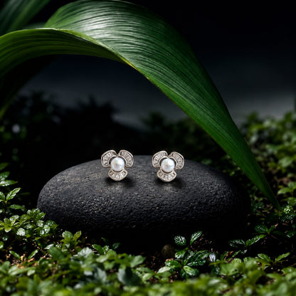 “Pair of silver-tone Pearl Arc Stud Earrings displayed on a black stone among deep green leaves, featuring white pearl centres and three baguette-stone arcs