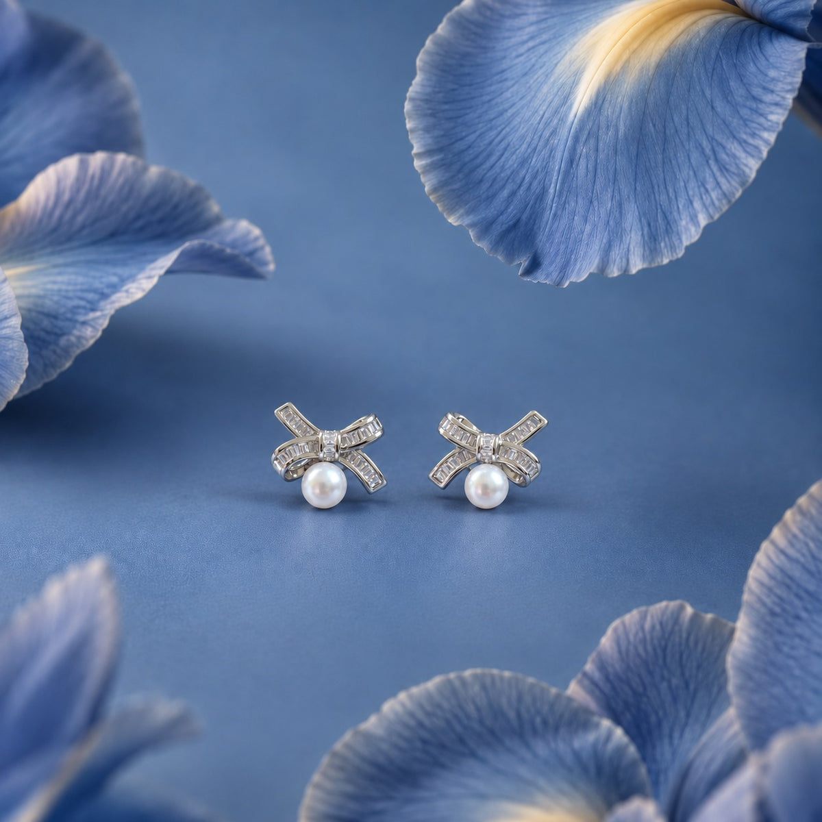 Pair of silver-tone Bowline Pearl Stud Earrings featuring zircon-set ribbon bows and round white pearl drops displayed against a soft blue floral setting