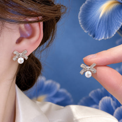 Woman wearing a silver-tone bow and pearl stud earring while holding the matching earring, showing the clear baguette zircon ribbon design and white pearl drop.
