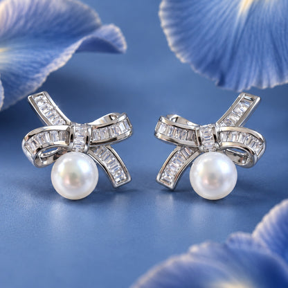 Close-up of silver bow stud earrings with clear baguette zircon detailing and luminous white pearls suspended beneath each bow.