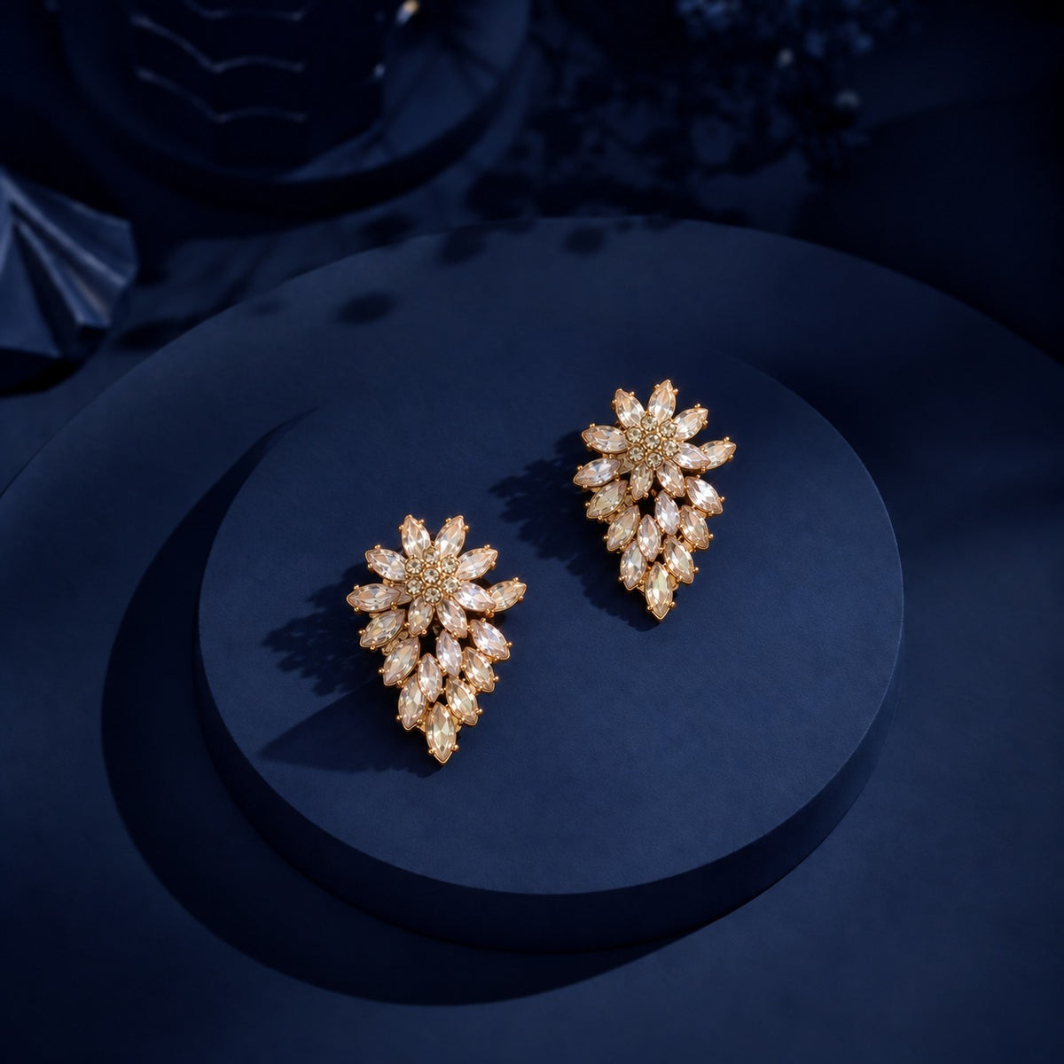 Pair of gold-tone Gilded Laurel Drop Earrings with clustered clear marquise stones displayed on a dark navy circular jewellery stand