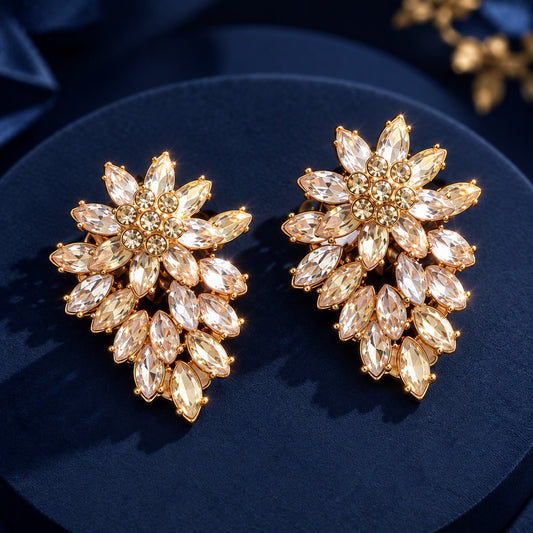 Close-up of Gilded Laurel Drop Earrings in gold-tone metal, featuring layered marquise-cut clear stones arranged in a floral tapered design on deep navy fabric