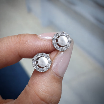 Close-up of Pearl Circuit Stud Earrings held between the fingers, showing white pearl centres, open circular construction and individually set clear stones