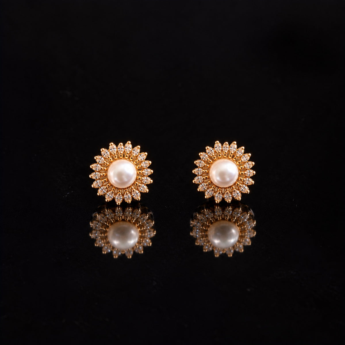 Pair of gold-tone Pearl Sunray Stud Earrings with round white pearl centres surrounded by radiating marquise-cut clear stones on a reflective black surfac