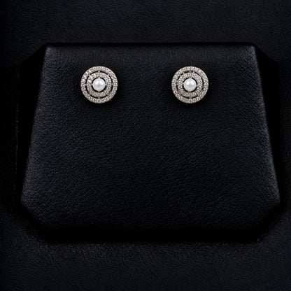 Pair of Pearl Axis Studs displayed on a black jewellery stand, featuring white pearl centres and double concentric zircon circles in silver-tone metal.