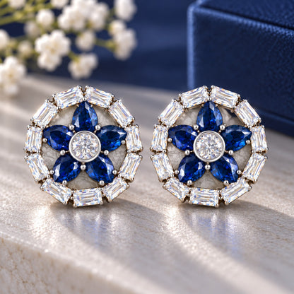 Petal Circuit Stud Earrings featuring five navy blue pear-cut coloured gemstone petals, a round clear zircon centre and a circular border of clear baguette stones in silver-tone metal.”